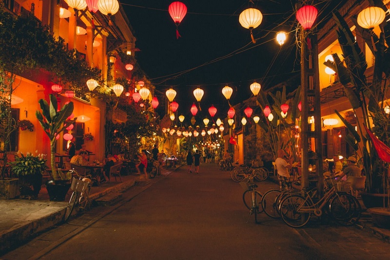 Lantern-lit street corner with tourists enjoying steaming bowls of noodles in Hoi An Ancient Town.